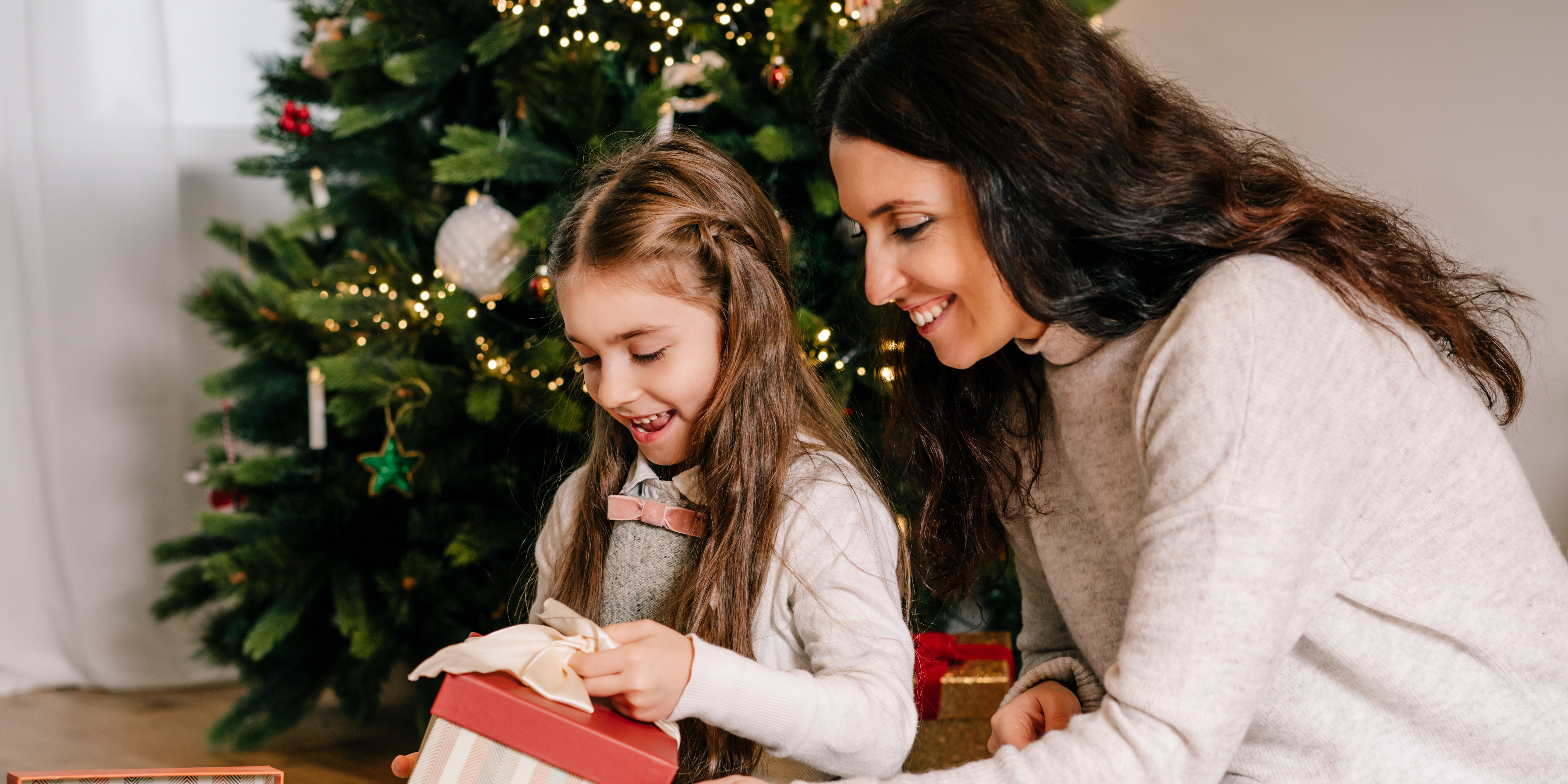 Mother and daughter sitting by a christmas tree looking at a gift box. 