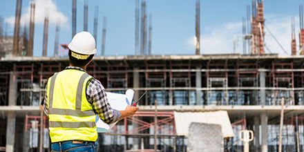 man wearing yellow vest and hard hat looking at building plans in front of job site