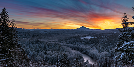 Horizon view of snowy forested trees with river, mountain and sunset.